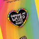 Katie Abey heart-shaped enamel pin displayed on a gradient background card. The black heart is encircled with rainbow-coloured petals and bears the message 'Following My Weird Little Heart'. The card also includes a playful declaration at the top encouraging the wearer to embrace their unique weirdness.