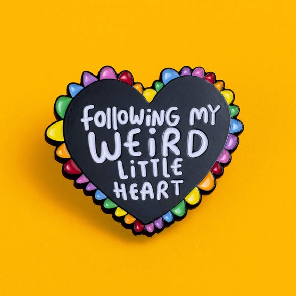 Close-up of a Katie Abey enamel pin featuring a black heart with rainbow-coloured petals around the edge. The heart is inscribed with the whimsical phrase 'Following My Weird Little Heart' in white, positioned against a solid yellow background