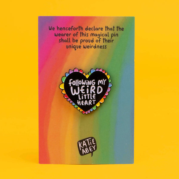 Katie Abey heart-shaped enamel pin displayed on a gradient background card. The black heart is encircled with rainbow-coloured petals and bears the message 'Following My Weird Little Heart'. The card also includes a playful declaration at the top encouraging the wearer to embrace their unique weirdness.