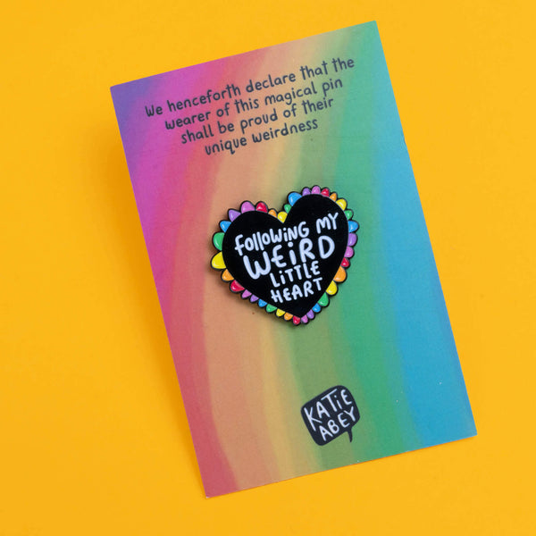 Katie Abey heart-shaped enamel pin displayed on a gradient background card. The black heart is encircled with rainbow-coloured petals and bears the message 'Following My Weird Little Heart'. The card also includes a playful declaration at the top encouraging the wearer to embrace their unique weirdness.