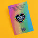 Katie Abey heart-shaped enamel pin displayed on a gradient background card. The black heart is encircled with rainbow-coloured petals and bears the message 'Following My Weird Little Heart'. The card also includes a playful declaration at the top encouraging the wearer to embrace their unique weirdness.