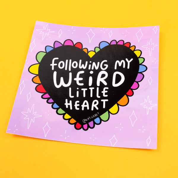 Katie Abey vinyl sticker displaying a vibrant rainbow-coloured heart, bordered with colourful petals. The heart bears the phrase 'Following My Weird Little Heart' in bold white letters against a black background, set against a soft lilac backdrop adorned with small diamond shapes.