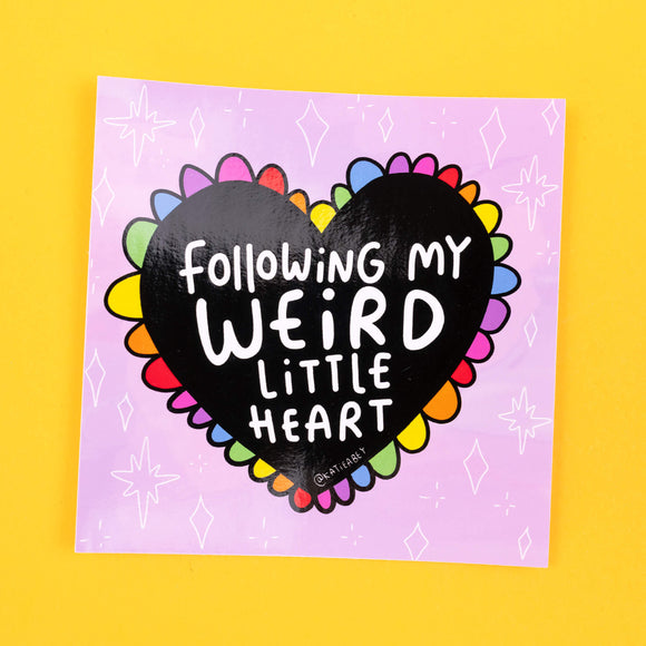 Katie Abey vinyl sticker displaying a vibrant rainbow-coloured heart, bordered with colourful petals. The heart bears the phrase 'Following My Weird Little Heart' in bold white letters against a black background, set against a soft lilac backdrop adorned with small diamond shapes.