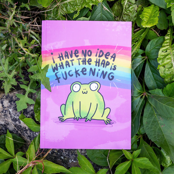 A purple notebook with an illustation of a cute smiling frog on the front with a rainbow above it and text that reads 'I have no idea what the hap is fuckening'. The notebook is placed amongst green leaves. Designed by Katie Abey in the UK.