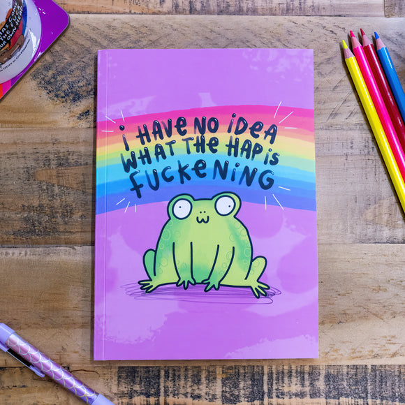 A purple notebook with an illustation of a cute smiling frog on the front with a rainbow above it and text that reads 'I have no idea what the hap is fuckening'. The notebook is on a wooden table amongst pens and pencils. Designed by Katie Abey in the UK.