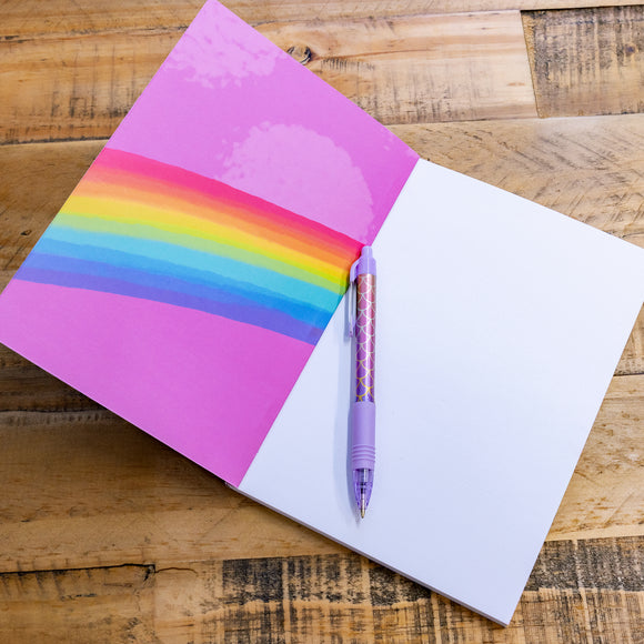 The I Have No Idea Frog Notebook of Necessity is open to reveal the first blank page with the purple backside of the front cover with a rainbow going across it. The notebook is on a wooden table with a pen laying on the blank page.