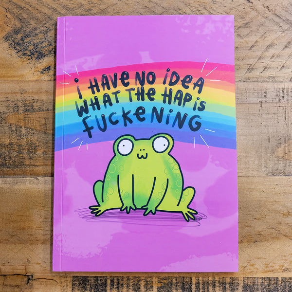 A purple notebook with an illustation of a cute smiling frog on the front with a rainbow above it and text that reads 'I have no idea what the hap is fuckening'. The notebook is on a wooden table. Designed by Katie Abey in the UK.