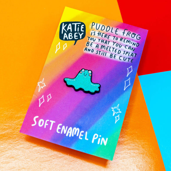 Puddle Frog Pin. A sparkly green frog shaped soft enamel pin with round white eyes, pink cheeks and cute smile. The pin is attached to rainbow coloured card with Katie Abey logo and writing that reads 'puddle frog is here to remind you that you can be a melted splat and still be cute'.