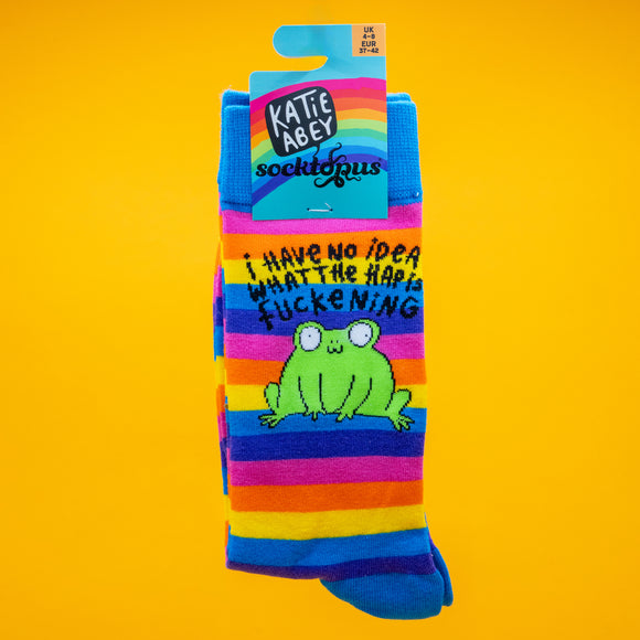 Bright rainbow striped Katie Abey socks featuring a green frog character and the phrase I Have No Idea What The Happ F*ckening. The socks are displayed against a bold yellow background. Fun, colourful novelty frog socks by illustrator Katie Abey encouraging you to embrace your weirdness.