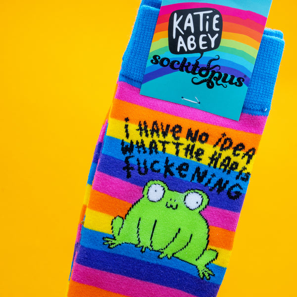 Close up of rainbow striped Katie Abey socks with a cute green frog and the phrase I Have No Idea What The Happ F*ckening. The socks are set against a vibrant yellow background. Colourful novelty frog socks by illustrator Katie Abey designed to spread joy and celebrate weirdness.