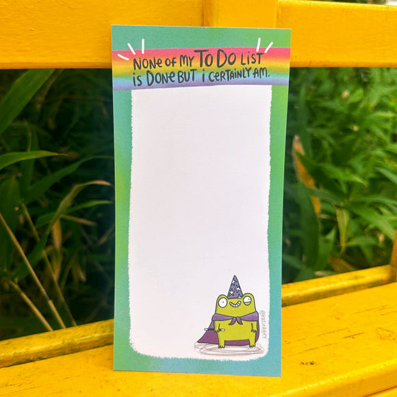 Katie Abey DL size notepad with the slogan "None Of My To Do List Is Done But I Certainly Am" at the top in a rainbow strip. The quirky pad has 50 tear-off sheets with a green border and a cartoon frog wizard illustration at the bottom. Shown outdoors against an outdoor yellow bench background. Fun stationery from indie illustrator Katie Abey, perfect for making to-do lists more magical.