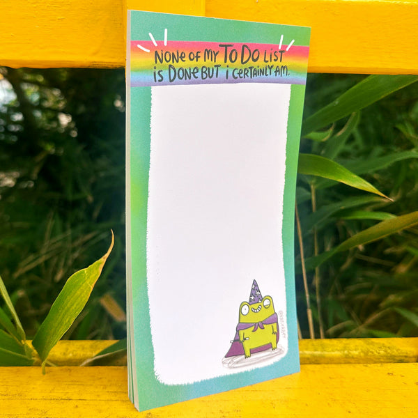 Katie Abey "None Of My To Do List Is Done But I Certainly Am" frog notepad shown propped outdoors against a yellow wooden bench. The DL size tear-off pad has a rainbow header with the funny slogan, a green border, and a cute illustrated frog in a wizard hat and cape at the bottom corner. Quirky stationery gift for list lovers from independent illustrator Katie Abey.