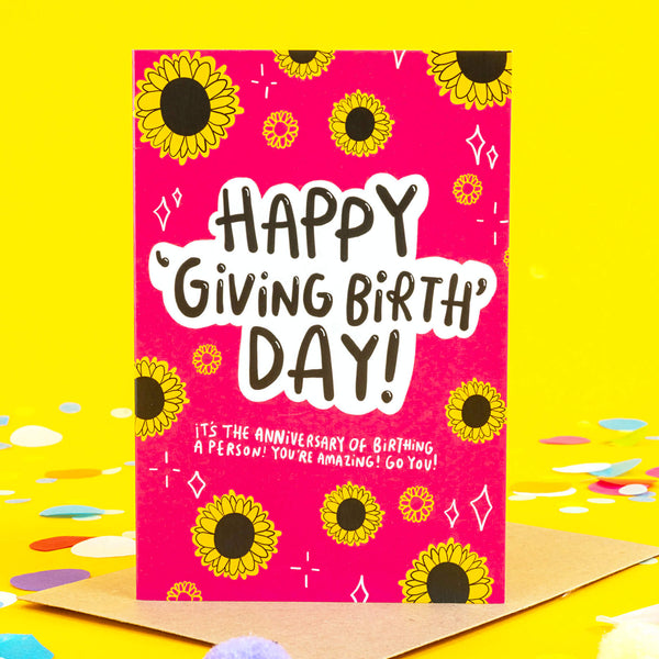 Happy giving birth day greeting card designed by Katie Abey and printed in the UK. The card has a bright pink front with various sunflowers and white sparkles around the border with text in the centre reading 'happy 'giving birth' day! it's the anniversary of birthing a person! you're amazing! go you!' The card is laying on a yellow background.