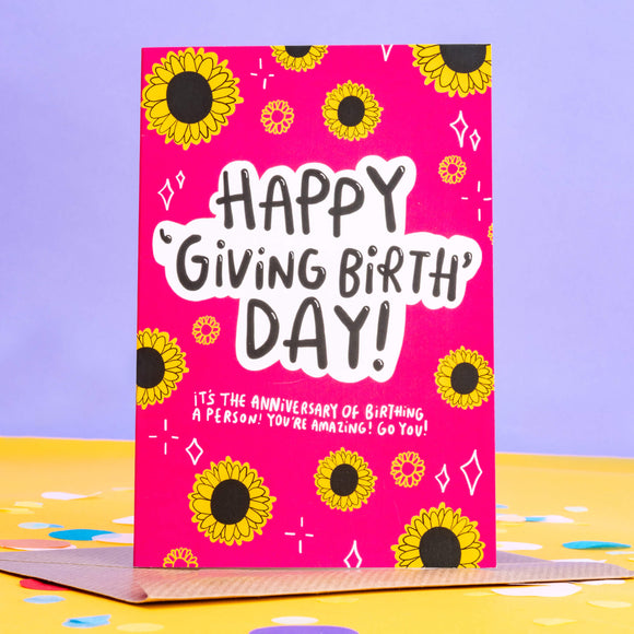 Happy giving birth day greeting card designed by Katie Abey and printed in the UK. The card has a bright pink front with various sunflowers and white sparkles around the border with text in the centre reading 'happy 'giving birth' day! it's the anniversary of birthing a person! you're amazing! go you!' The card is on a lilac and yellow background.