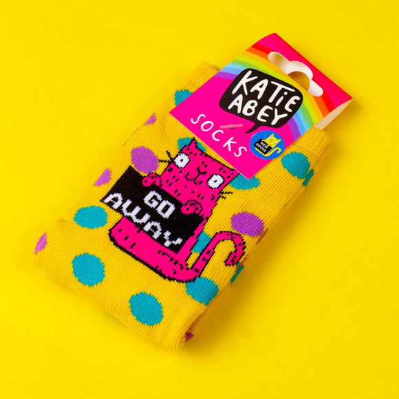 Yellow socks with blue and purple polka dots and pink cat illustration holding a black sign that reads 'go away'. There is a pink cardboard label attached to the socks with a rainbow and Katie Abey logo on. The socks are on a bright yellow background.