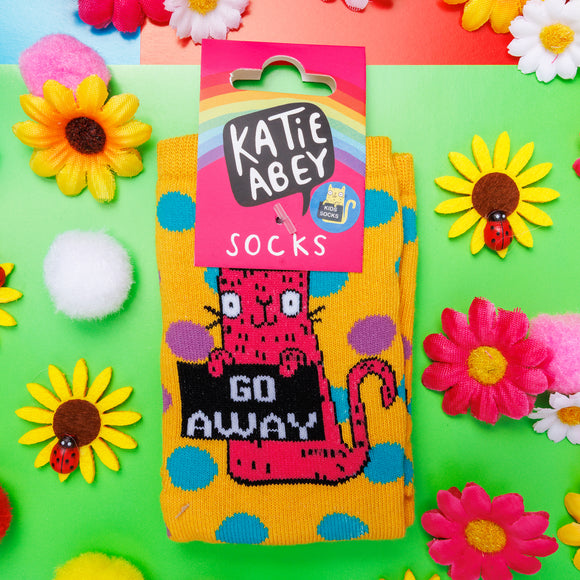 Yellow socks with blue and purple polka dots and pink cat illustration holding a black sign that reads 'go away'. There is a pink cardboard label attached to the socks with a rainbow and Katie Abey logo on. The socks are lying on a green background with flowers and ladybugs.