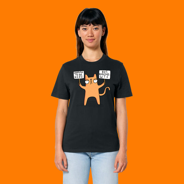 Front view of a model wearing the Katie Abey Grateful To Be Here But Respectfully WTF black graphic t shirt. Funny cartoon cat illustration by Katie Abey holding humorous protest signs. Alternative unisex tee styled with jeans against a bright orange background.