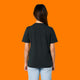 Back view of a model wearing the Katie Abey Grateful To Be Here But Respectfully WTF black graphic t shirt. Unisex organic cotton tee with a plain back and classic crew neck, styled with light denim jeans against an orange studio background.