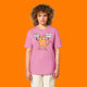 Front view of a model wearing the Katie Abey Grateful To Be Here But Respectfully WTF bubble pink graphic t shirt. Features a quirky orange cartoon cat holding signs reading “Grateful To Be Here” and “But Respectfully… WTF”, illustrated by Katie Abey.
