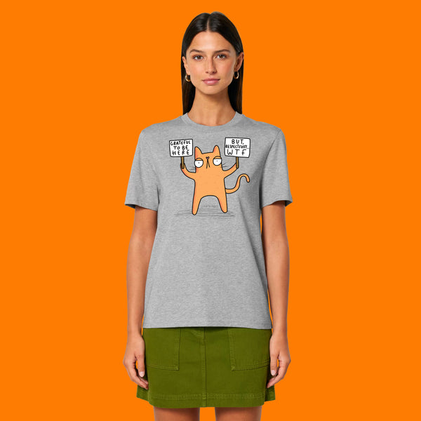 Front view of a model wearing the Katie Abey Grateful To Be Here But Respectfully WTF heather grey unisex adults t shirt. Funny cartoon cat illustration by Katie Abey with “Grateful To Be Here But Respectfully WTF” slogan. Styled with a green skirt on an orange background.
