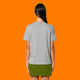 Back view of a model wearing the Katie Abey Grateful To Be Here But Respectfully WTF heather grey adults t shirt. Unisex fit organic cotton graphic tee with plain back, styled with a green skirt against an orange studio background.
