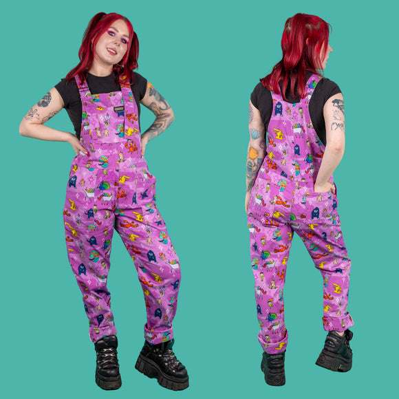 Happiness Enchanters x Run & Fly Word Spells Stretch Twill Dungarees worn by Flo with a black short sleeve tshirt and black platform trainers on a teal blue background. She has red short hair in top bunches with neon pink eye makeup and tattoos. Two angles show flo facing forward smiling with both hands on her hips and facing away with her hand in the back pocket. Pastel pink & light purple dungarees feature various mythical creatures such as mermaids, dragons & unicorns with white & pink sparkles.