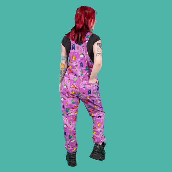The Run & Fly x Happiness Enchanters Word Spells Stretch Twill Dungarees being worn by Flo with a black short sleeve tshirt and black platform trainers in front of a teal blue background. She has red short hair in top bunches with neon pink eye makeup and tattoos. She is facing away from the camera with one leg bent and one hand in the back pocket. The pastel pink and light purple dungarees feature various mythical creatures such as mermaids, dragons and unicorns with white and pink sparkles.