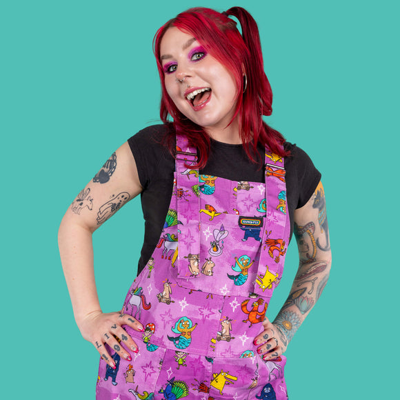 The Happiness Enchanters x Run & Fly Word Spells Stretch Twill Dungarees being worn by Flo with a black short sleeve tshirt and black platform trainers in front of a teal blue background. She has red short hair in top bunches with neon pink eye makeup and tattoos. She is facing forward with both hands on her hips smiling. The pastel pink and light purple dungarees feature various mythical creatures such as mermaids, dragons and unicorns with white and pink sparkles.