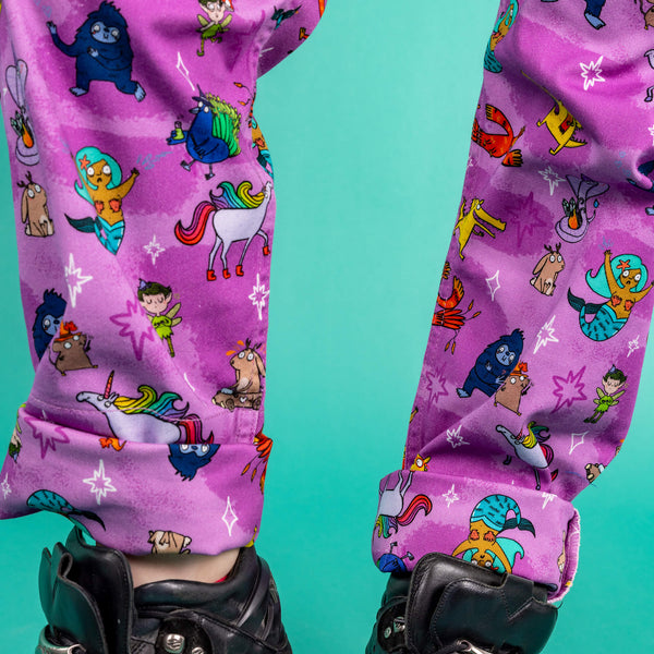 The Run & Fly x Happiness Enchanters Word Spells Stretch Twill Dungarees being worn by Flo with black platform trainers in front of a teal blue background. The photo is cropped in on the lower legs to show the matching turn ups. The pastel pink and light purple dungarees feature various mythical creatures such as mermaids, dragons and unicorns with white and pink sparkles.