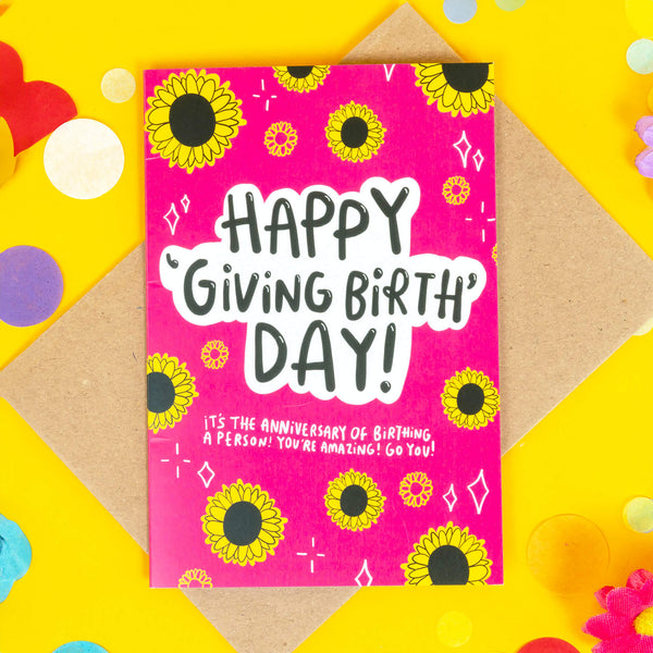 Happy giving birth day greeting card designed by Katie Abey and printed in the UK. The card has a bright pink front with various sunflowers and white sparkles around the border with text in the centre reading 'happy 'giving birth' day! it's the anniversary of birthing a person! you're amazing! go you!' The card is laying on a yellow background.
