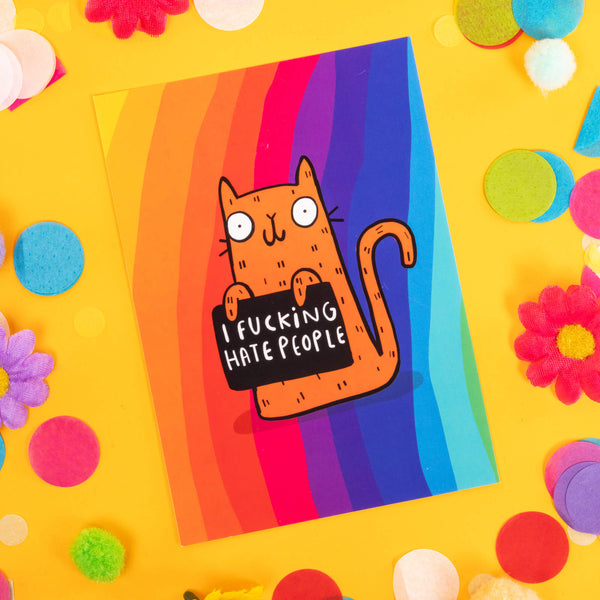 A6 greeting postcard featuring smiley illustrated orange and black cat holding sweary sign, on rainbow background. Designed by Katie Abey in the UK