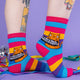 a model with tattooed legs wearing Katie Abey I fucking hate people socks with an orange cat holding a sign. They are rainbow striped and lovely and vibrant. The model is stood on a yellow floor with balloons, confetti and ribbons