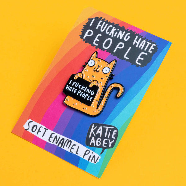 Katie Abey hand drawn illustration enamel pin featuring a bright orange cat with wide, bulging eyes and a mischievous expression, holding a black sign with the bold statement 'I Fucking Hate People'. The pin is attached to its rainbow backing card with Katie Abey logo at the bottom.