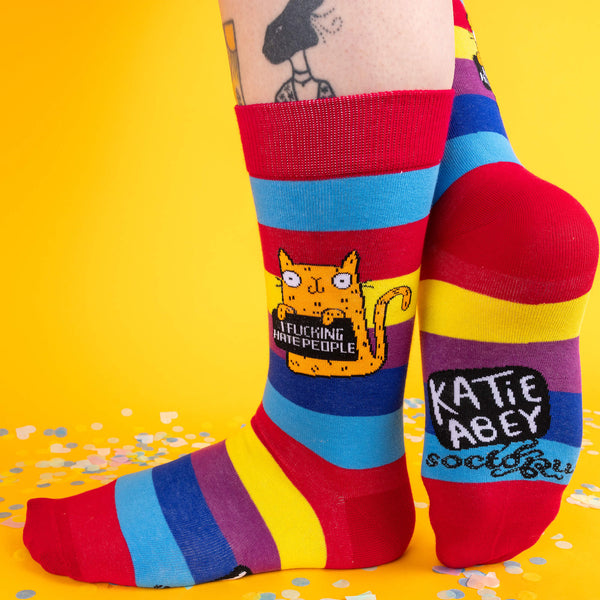 Close-up side view of Katie Abey’s “I F*cking Hate People” rainbow socks. Bold stripes in red, yellow, blue, purple, and green showcase an orange cat holding a sign with the humorous phrase. Perfect for anyone with a sassy sense of style.