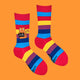 A flat lay of Katie Abey's rainbow-striped socks with an illustrated orange cat holding a cheeky sign reading, "I F***ing Hate People." Fun and funky, these socks make a statement while embracing colourful self-expression.