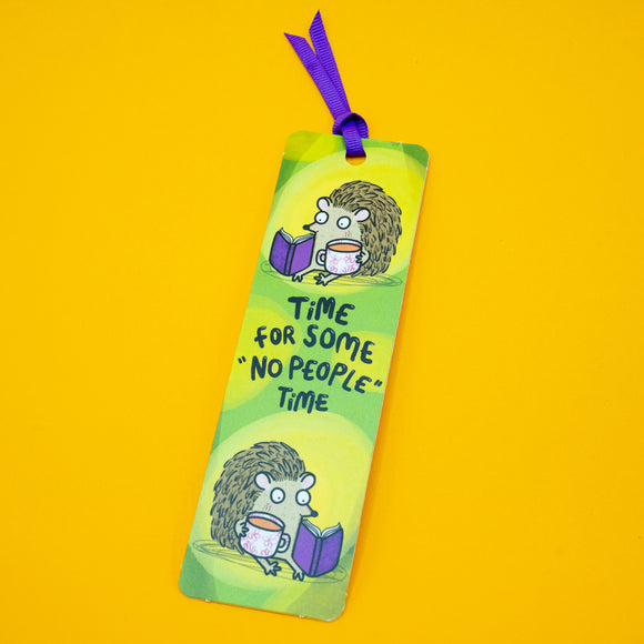 Angled view of Katie Abey hedgehog bookmark with purple ribbon on yellow background. Features hedgehog reading illustration and “Time for some ‘No people’ time” quote. Fun, colourful bookmark gift for readers, introverts and fans of cute animal designs.