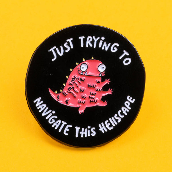 Close-up of a Katie Abey enamel pin with a red, quirky, multi-limbed creature and the phrase 'Just Trying to Navigate This Hellscape'. The pin is set against a bright yellow background, highlighting its bold design.