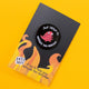 Katie Abey enamel pin featuring a quirky red creature with multiple eyes and limbs, surrounded by the text 'Just Trying to Navigate This Hellscape' on a black circular background. The pin is placed on a dark background with a hint of yellow in the corner.