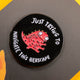 Katie Abey enamel pin featuring a quirky red creature with multiple eyes and limbs, surrounded by the text 'Just Trying to Navigate This Hellscape' on a black circular background. The pin is placed on a dark background with a hint of yellow in the corner.