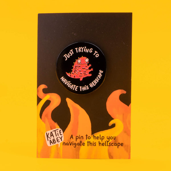 Katie Abey enamel pin on its original packaging card, showing a red creature with multiple eyes and the text 'Just Trying to Navigate This Hellscape'. The card has a yellow flame design and the phrase 'A pin to help you navigate this hellscape', capturing the brand's unique, playful style.