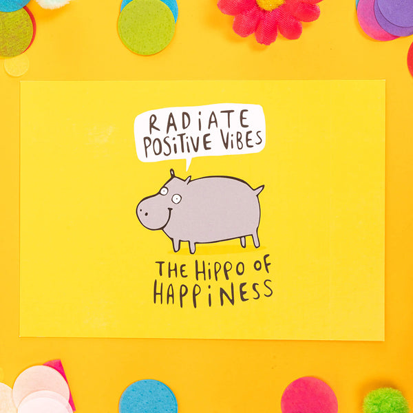 yellow postcard of the hippo of happiness saying radiate positive vibes illustrated by Katie Abey. It is on a yellow background with confetti, fake daises and pom poms are in bright colours
