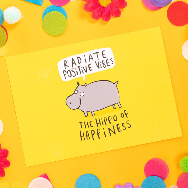 yellow postcard of the hippo of happiness saying radiate positive vibes illustrated by Katie Abey. It is on a yellow background with confetti, fake daises and pom poms are in bright colours