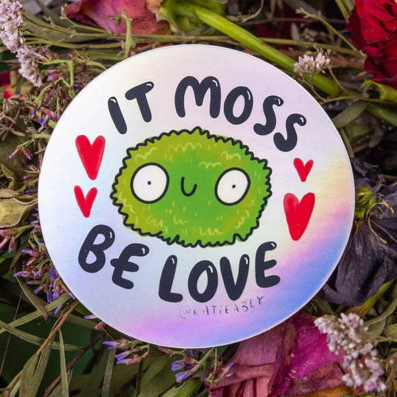 The It Moss be Love Holographic Vinyl Sticker on purple, red, pink and green foliage. The silver rainbow holographic circular sticker has a smiling ball of moss with red hearts and black text reading 'it moss be love' surrounding. 