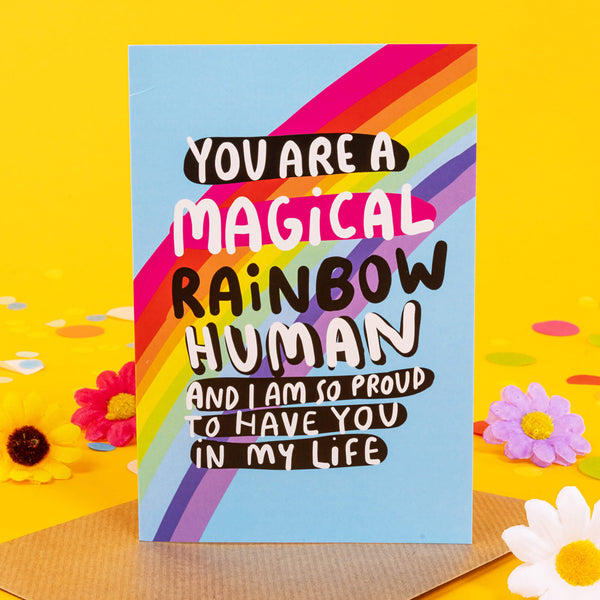 A6 Greetings Card that reads 'You Are a Magical Rainbow Human and I am so proud to have you in my life' with black white pink writing on blue background with rainbow illustration. Designed by Katie Abey in the UK