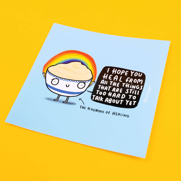 Katie Abey vinyl sticker featuring a cute, cartoon hummus character, with a rainbow arching above its head. The character holds a speech bubble that says, 'I Hope You Heal From All The Things That Are Still Too Hard To Talk About Yet', with the label 'The Houmous of Healing' beneath.