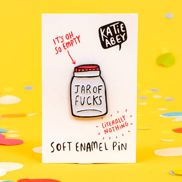 Katie Abey soft enamel pin featuring a whimsical design of a jar labelled 'Jar of Fucks' with text above it saying 'It's Oh So Empty' and text below it saying 'Literally Nothing'. The pin is displayed on a white card with the artist's name, Katie Abey, in a speech bubble at the top right. The background is a cheerful yellow with scattered colourful confetti, emphasising the humorous and light-hearted nature of the product.