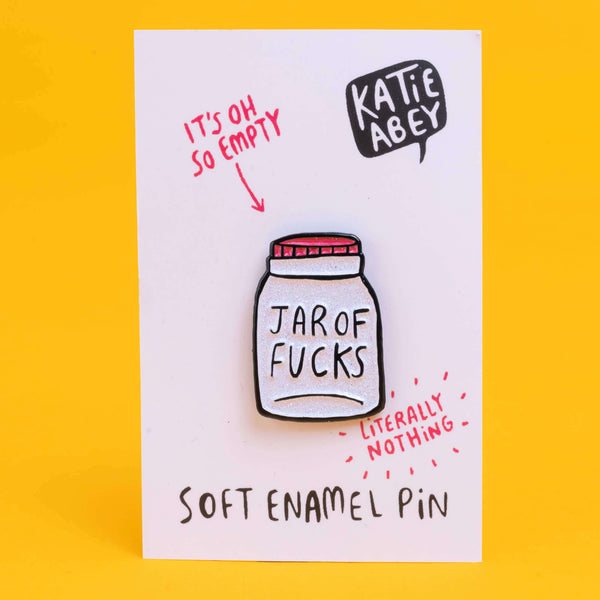 Katie Abey soft enamel pin featuring a whimsical design of a jar labelled 'Jar of Fucks' with text above it saying 'It's Oh So Empty' and text below it saying 'Literally Nothing'. The pin is displayed on a white card with the artist's name, Katie Abey, in a speech bubble at the top right.  The pin is sat against a yellow backdrop.