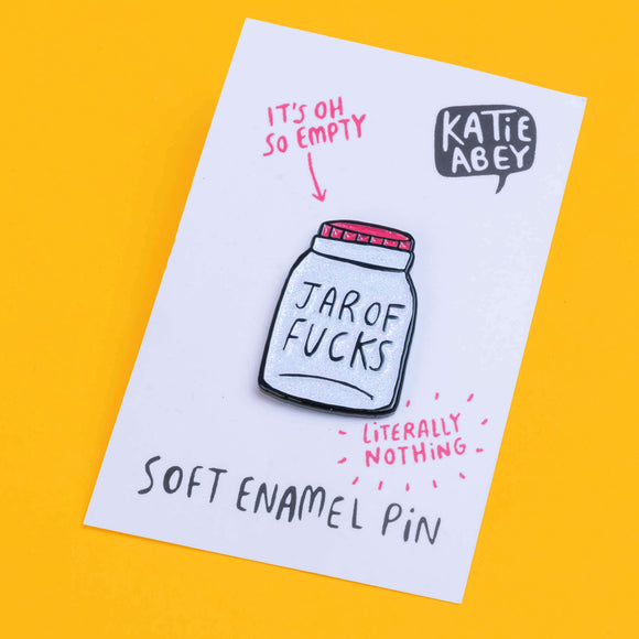 Katie Abey soft enamel pin featuring a whimsical design of a jar labelled 'Jar of Fucks' with text above it saying 'It's Oh So Empty' and text below it saying 'Literally Nothing'. The pin is displayed on a white card with the artist's name, Katie Abey, in a speech bubble at the top right.  The pin is sat against a yellow backdrop.