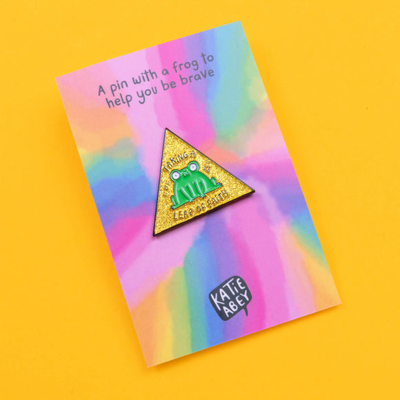 Katie Abey 'Leap of Faith' frog pin displayed on a rainbow background card. The pin features a quirky green frog on a glittery yellow triangle with the words 'Taking a Leap of Faith'. The card reads, 'A pin with a frog to help you be brave'.