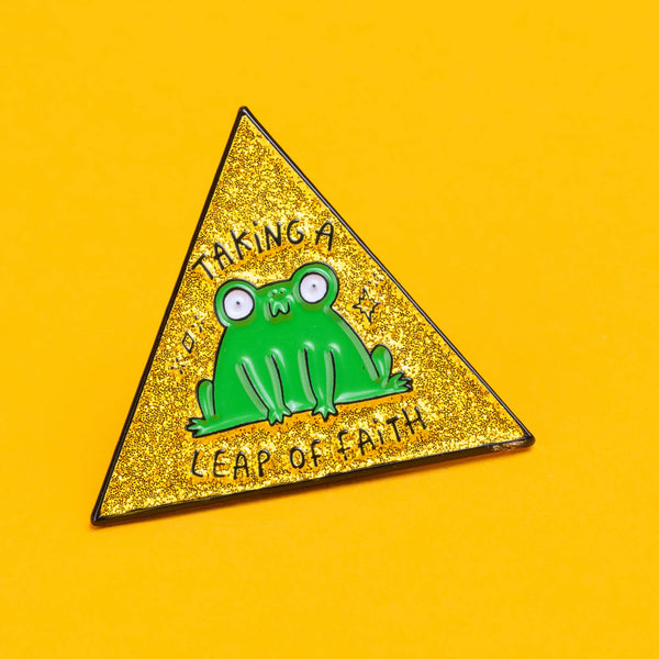 Close-up of the Katie Abey 'Leap of Faith' enamel pin featuring a quirky green frog on a yellow glittery triangle background. The words 'Taking a Leap of Faith' are written around the frog.